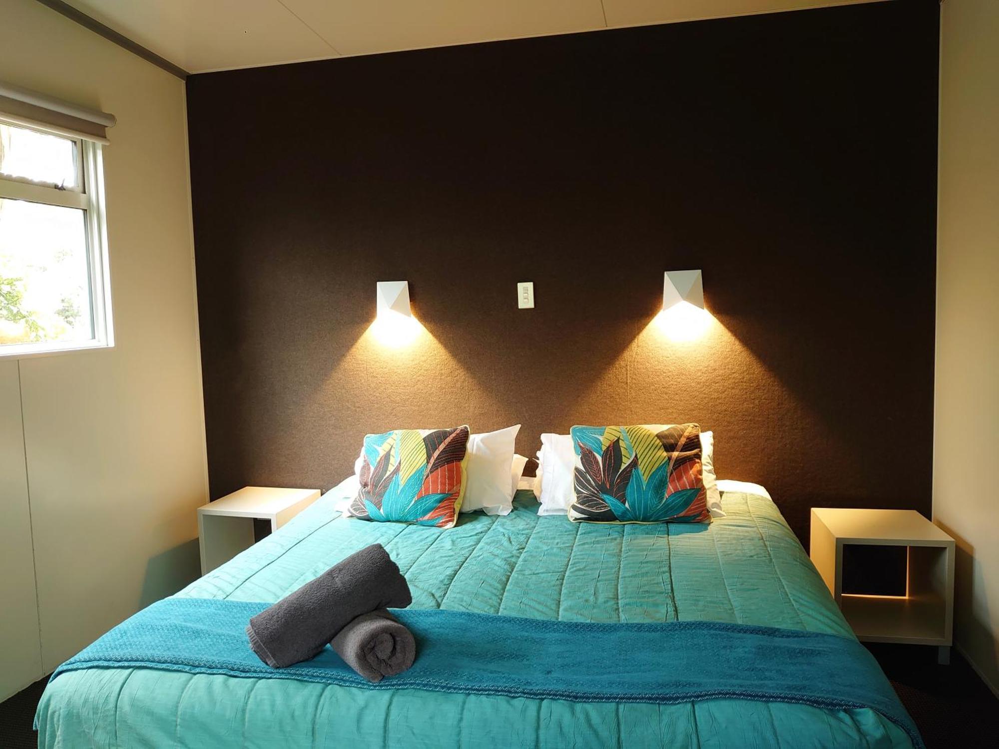 タウポ デブレッツ スパ リゾート エクステリア 写真 The image shows a bedroom with a modern and inviting layout. The focal point is a bed with a vibrant turquoise comforter, decorated with two colorful pillows featuring floral patterns. On either side of the bed, there are small nightstands with wall-