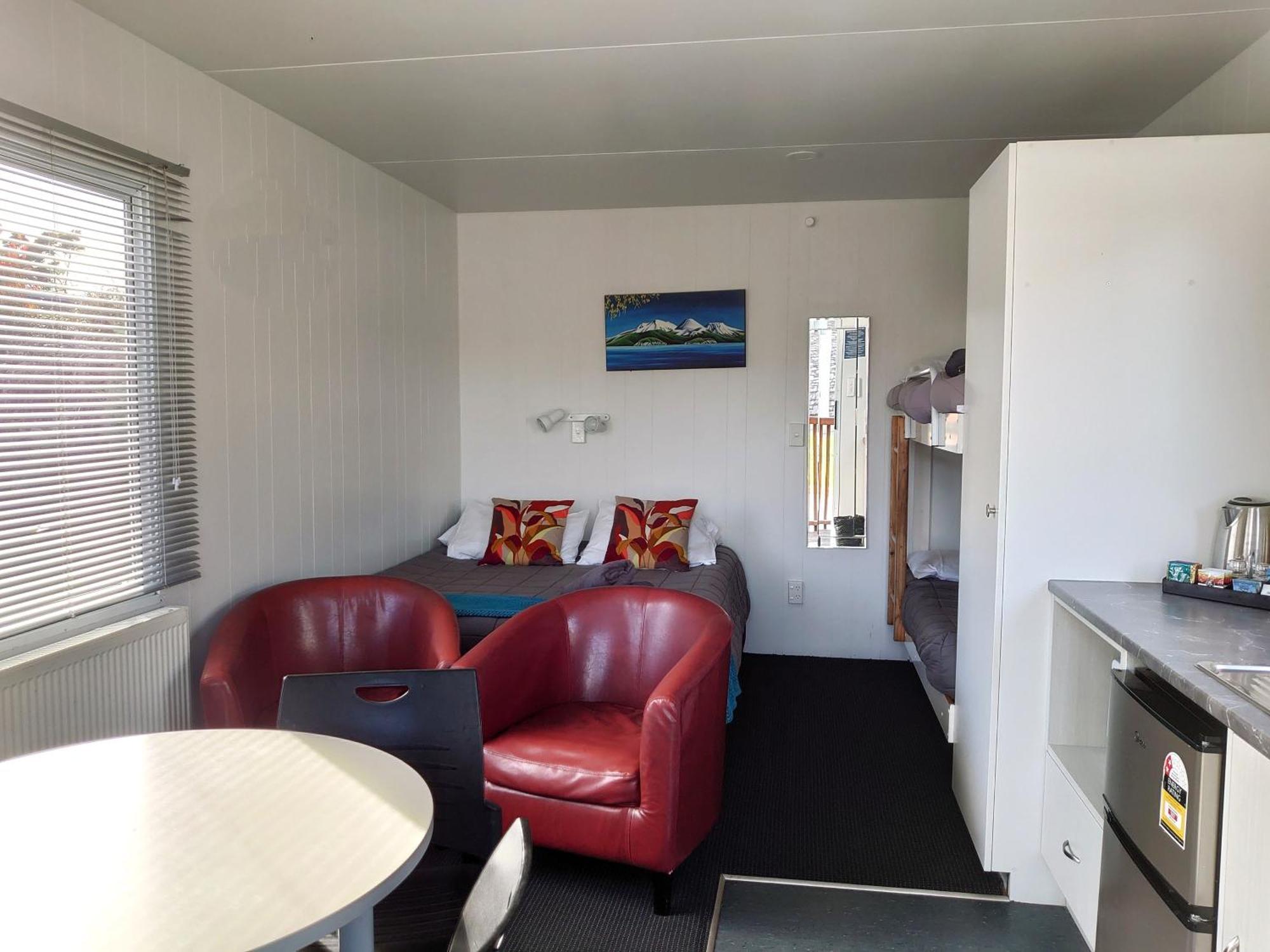 タウポ デブレッツ スパ リゾート エクステリア 写真 The photo shows the interior of a small living space, possibly a cabin or studio apartment. There are two red armchairs and a round table in the foreground. The wall opposite has a bed with colorful pillows and a small nightstand. To the side, there 